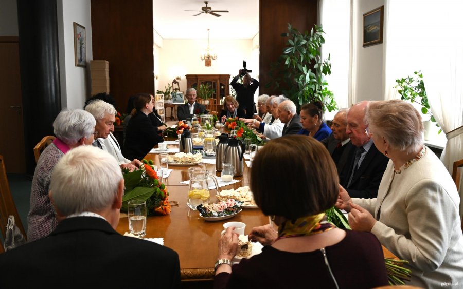 Grupa os&oacute;b przy stole, na stole kawa i ciasto