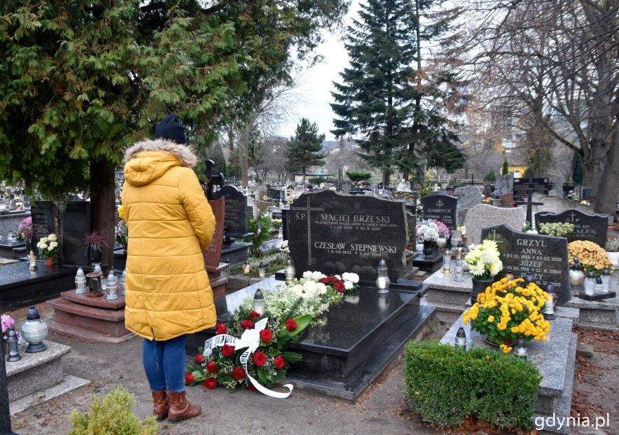 Na zdjęciu wieniec z czerwonych róż złożony na mogile z ciemną granitową tablicą z napisem Maciej Brzeski i Czesław Stępniewski, przed grobem stoi postać w żółtej kurtce