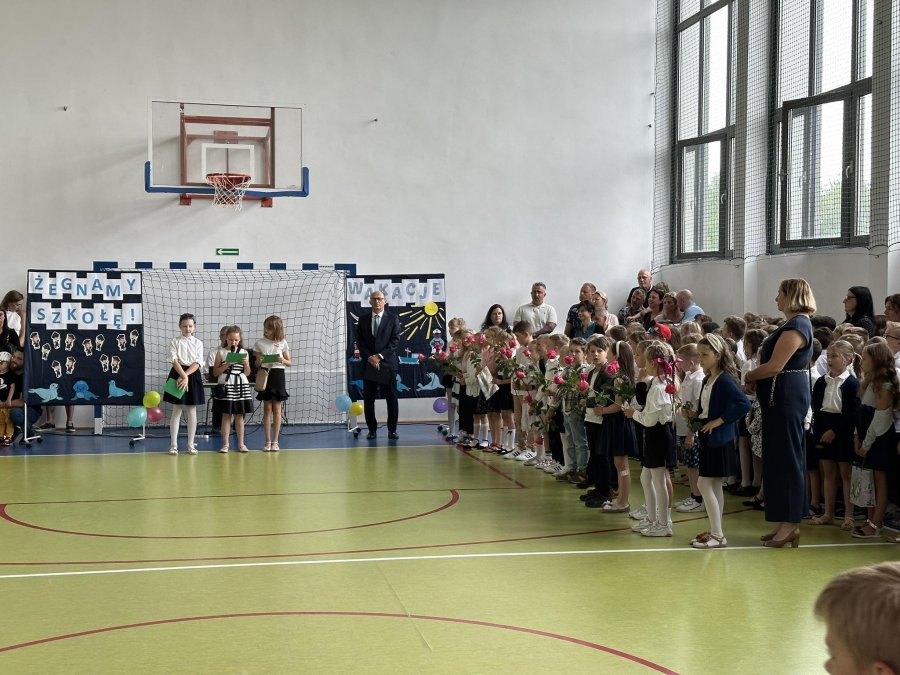 Uroczystość w hali sportowej z lewej strony dzieci z kwiatami