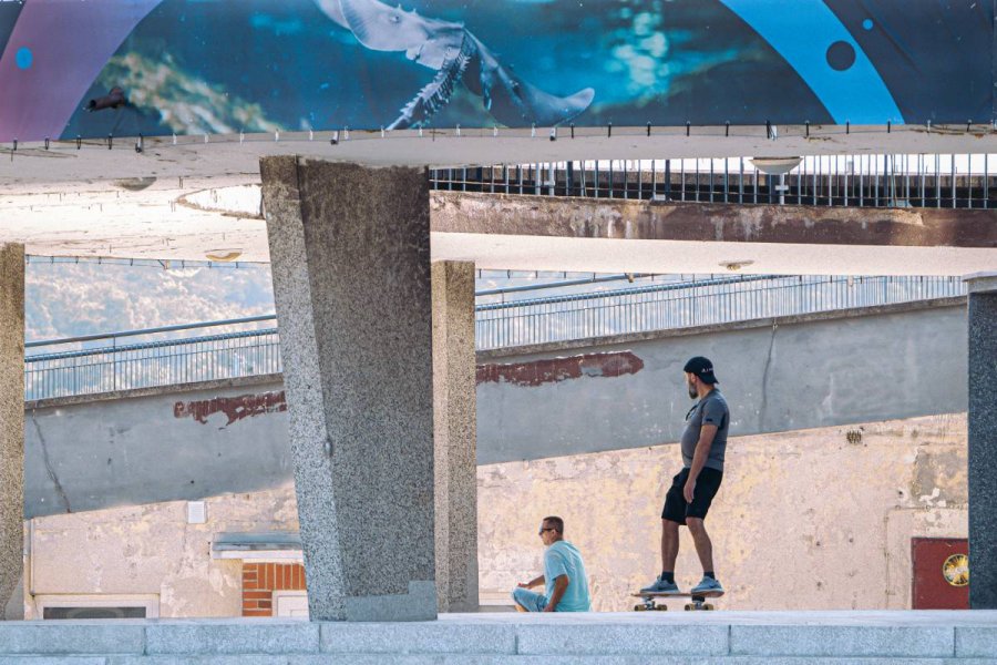 Mężczyzna jadący na deskorolce pod estakadą i mijający drugiego, siedzącego mężczyznę