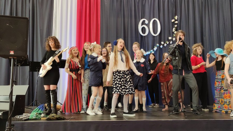 Na scenie grupa przebranych uczniów z instrumentami w tle kotara, ze spływającą z sufitu flagą Polski oraz gałązka z białymi kwiatami a nad nią liczba 60