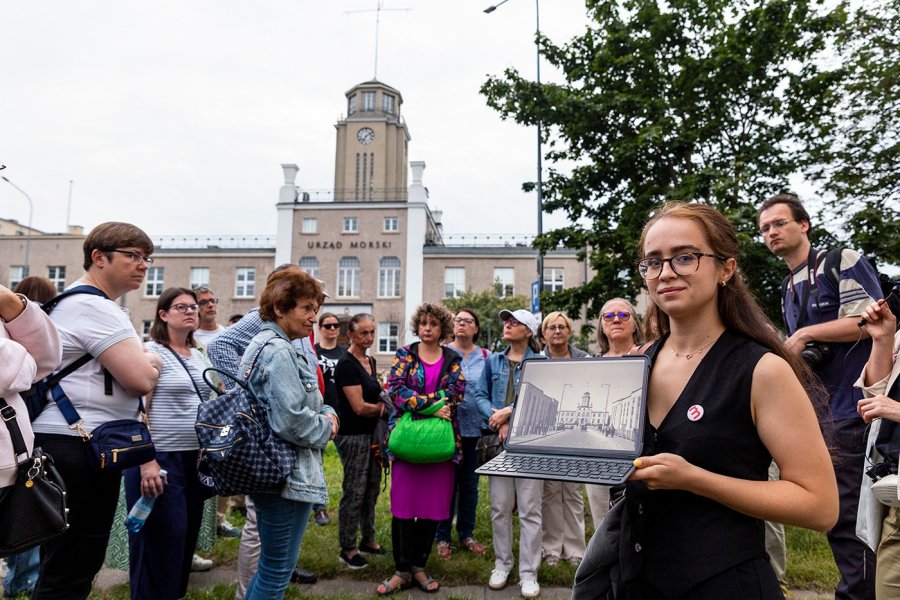Spacer otwarcia: ludzie przy Urzędzie Morskim, prowadząca dr Anna Orchowska z Wydziału Architektury PG trzyma otwarty tablet z historycznym zdj. tego obiektu