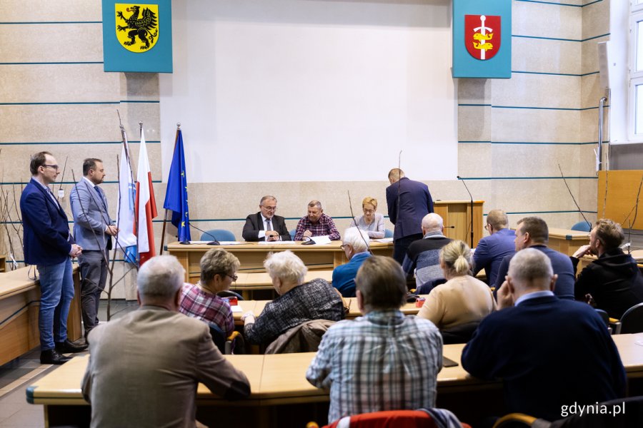 Na sali siedzą ludzie za stołami, przed nimi za stołem inni ludzie podpisują umowy.