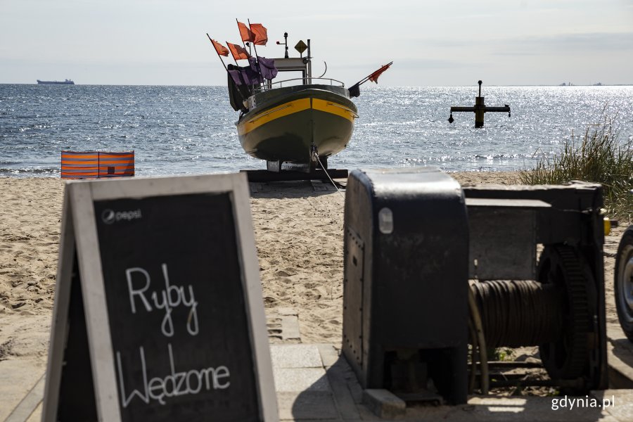 Plaża w Orłowie. Na pierwszym planie tablica z napisem "ryby wędzone", obok wyciągarka łodzi, w tle kuter rybacki na piasku i morze. Fot, Przemysław Kozłowski