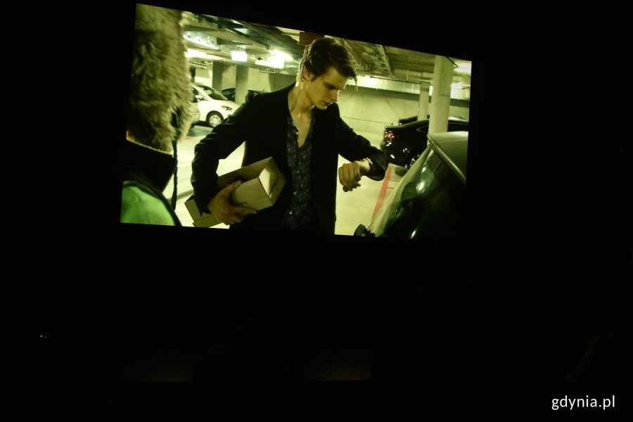 Kadr z filmu, który przygotowali studenci pierwszego roku reżyserii z okazji rozpoczęcia nowego roku akademickiego w Gdyńskiej Szkole Filmowej. W kadrze: chłopak w czarnej koszuli i marynarce stoi na parkingu obok samochodu, w ręku trzyma pudełko z butami // fot. Magdalena Czernek