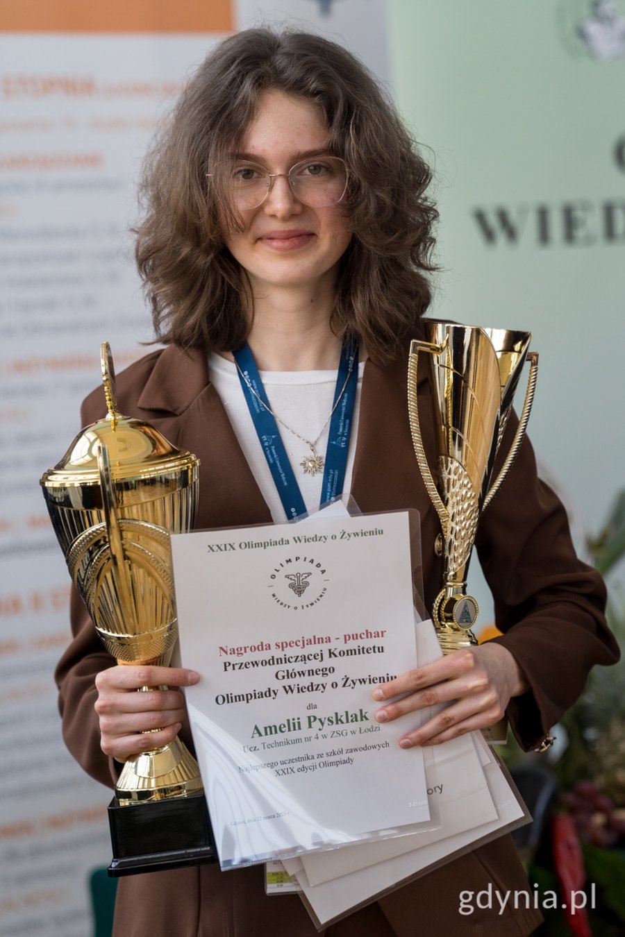 dziewczyna z kręconymi włosami do ramion i w okularach, ubrana w brązowa marynarkę, trzymająca 2 puchary oraz dyplomy 