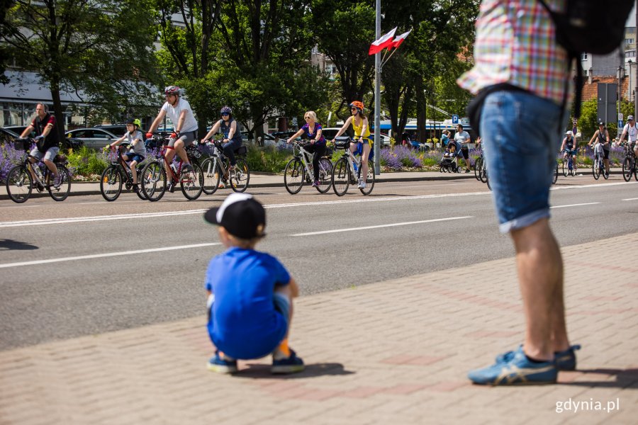 na zdjęciu mały chłopiec kuca i przygląda się rowerzystom jadącym ulicą