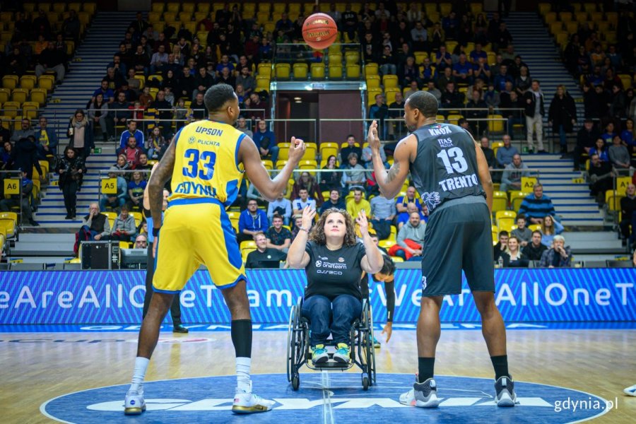 Koszykarze oraz kobieta na wózku inwalidzkim, która podrzuca do góry piłkę do koszykówki. 