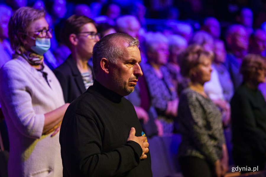 Mężczyzna w czarnym golfie trzyma rękę na piersi, w tle widzowie zgromadzenie w Teatrze Muzycznym wstali z miejsc podczas odgrywania hymnu Ukrainy // fot. Przemysław Kozłowski