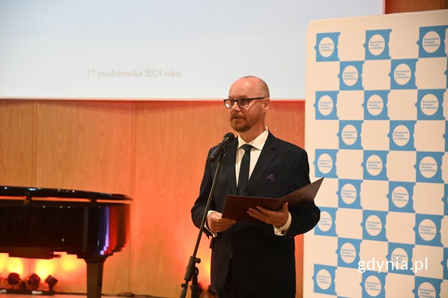 Mężczyzna elegancko ubrany i trzymający teczkę stoi na scenie przed mikrofonem