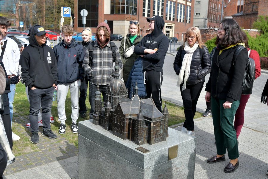 Na pierwszym planie na postumencie model synagogi ze spiżu, w tle grupa młodzieży na drugim planie budynki z czerwonej cegły. 