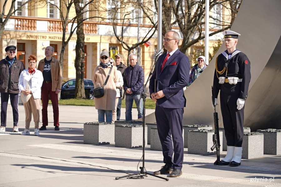 Wicewojewoda pomorski Emil Rojek przemawiający podczas uroczystości z okazji 80. rocznicy zakończenia walk o Gdynię. Za nim: marynarz z posterunku honorowego pod pomnikiem Polski Morskiej oraz grupa mieszkańców