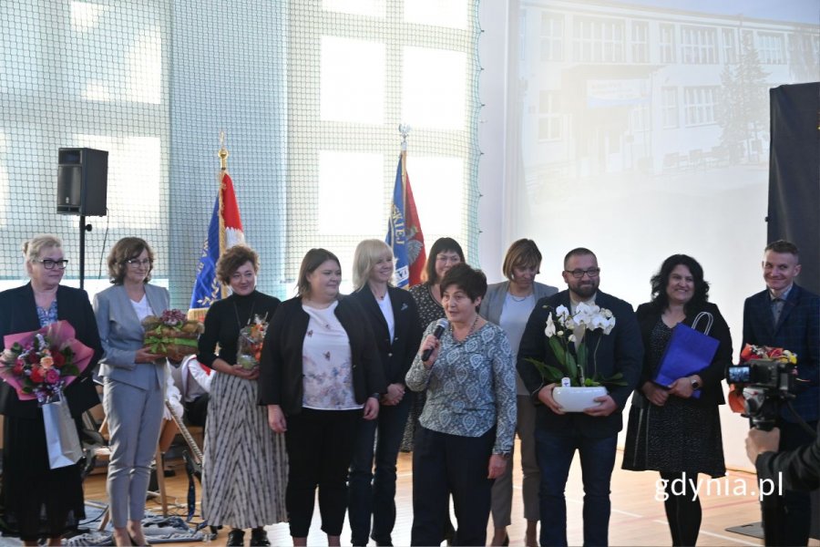 Stojąca kobieta mówiąca do mikrofonu w otoczeniu innych osób w szkolnej hali sportowej