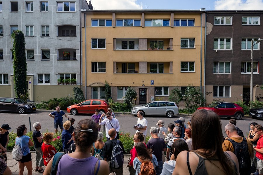 Uczestnicy archiwalnego Archi-Spaceru &bdquo;Wakacje z modernizmem&rdquo; słuchają przewodnika przed budynkiem mieszkalnym.
