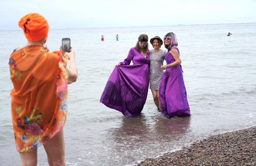 Kobieta w pomarańczowym stroju robi zdjęcie dw&oacute;m paniom w sukniach w morzu