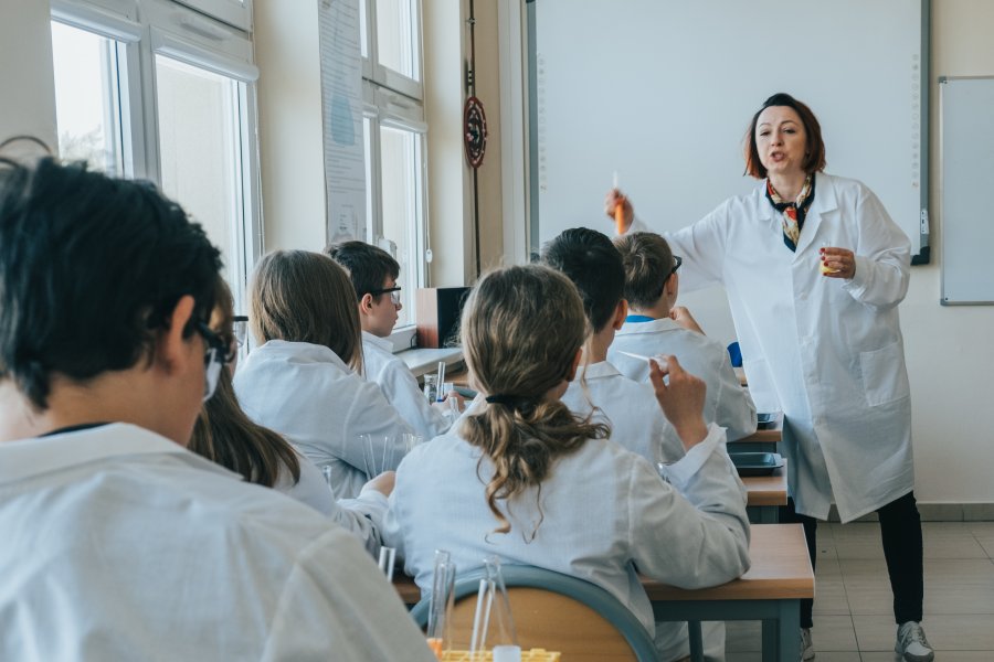 Nauczycielka w białym fartuchu, z uczniami, też w fartuchach w trakcie doświadczeń chemicznych
