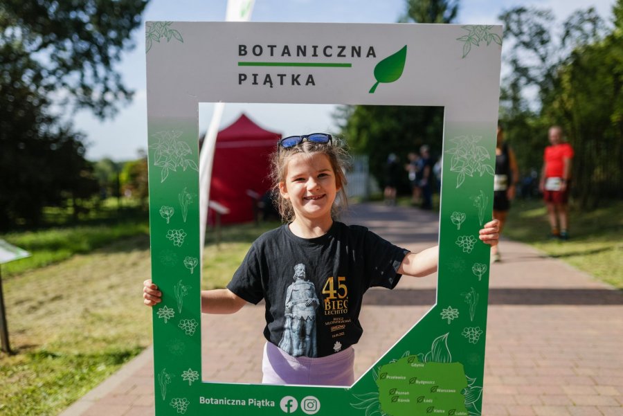 Uśmiechnięta dziewczynka trzymająca kartonową ramkę do zdjęć z napisem Botaniczna Piątka. 
