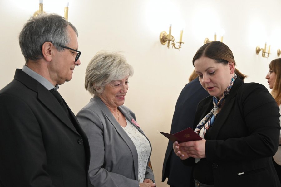 Starsi państwo i kobieta w gratanowej marynarce, kt&oacute;ra trzyma w ręce czerwoną książeczkę