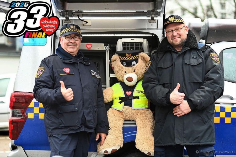 Na zdjęciu widać dwóch strażników miejskich w Gdyni podczas 33. finału Wielkiej Orkiestry Świątecznej Pomocy. Pomiędzy nimi maskotka.