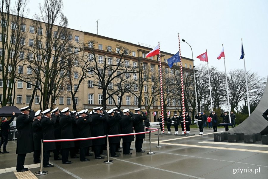 Flagi Polski i NATO wciągnięte na maszt podczas uroczystości z okazji 25. rocznicy wstąpienia Polski do NATO. Przed masztami salutujący przedstawiciele Marynarki Wojennej