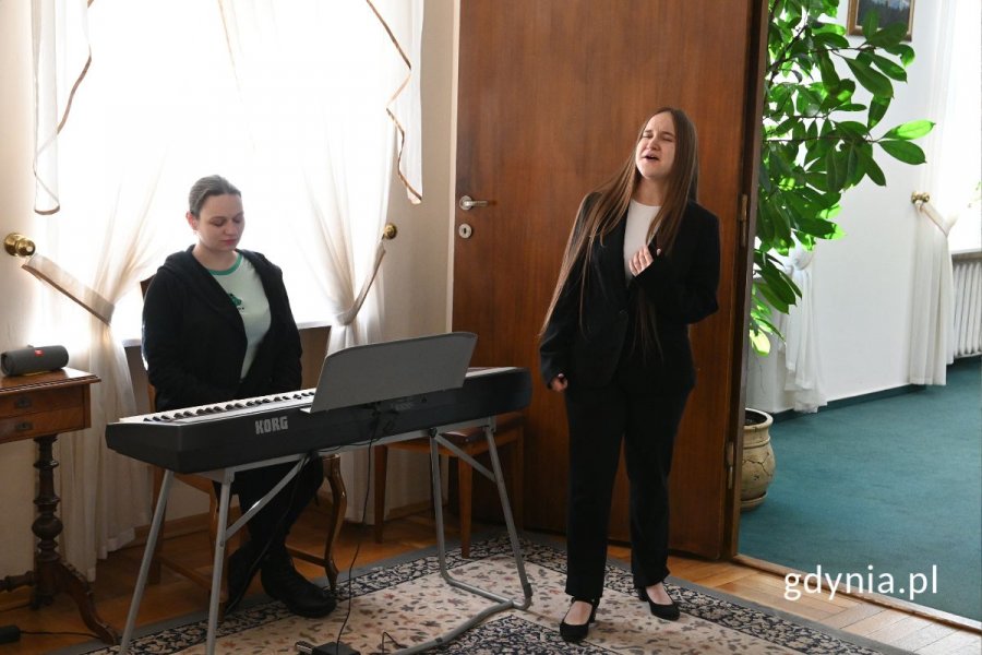 Elegancko ubrana kobieta stojąca i śpiewająca w eleganckiej sali, obok kobieta siedząca przy keyboardzie