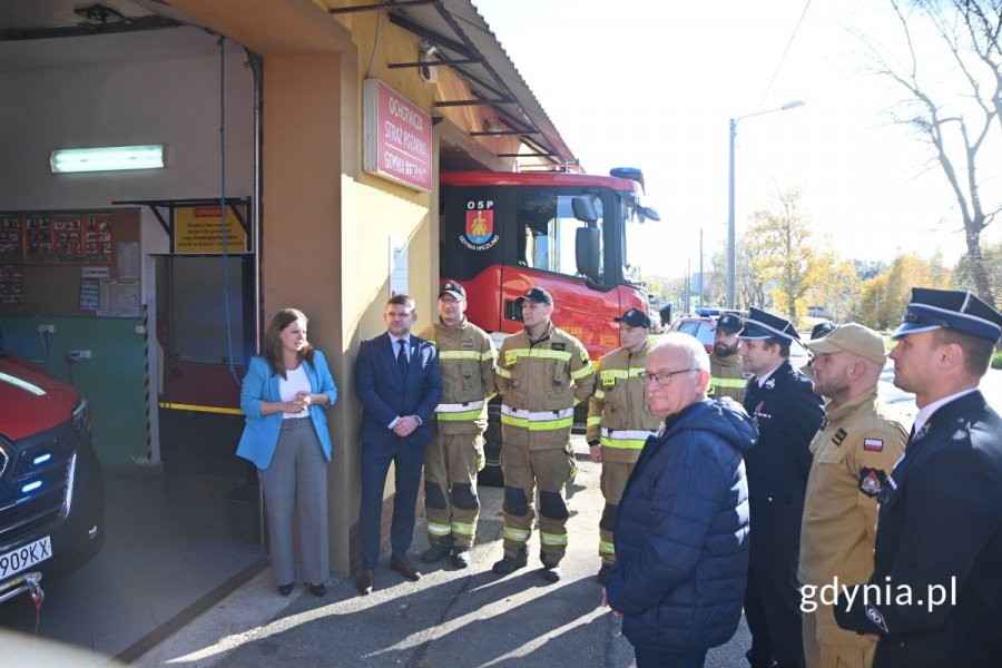 Strażacy, ratownicy i osoby w strojach cywilnych zgromadzone przed garażem, w którym stoi czerwony furgon