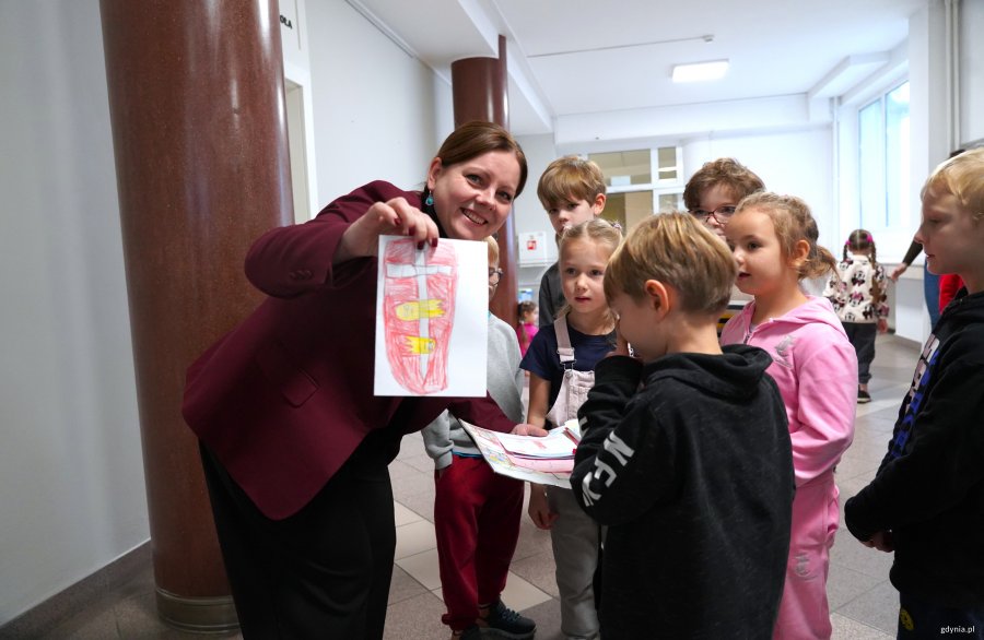 Kobieta w bordowej marynarce pokazuje dziecięcy rysunek, obok dzieci