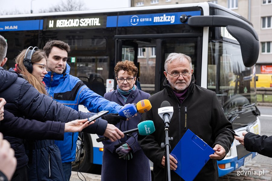 Mężczyzna mówi do mikrofonów w otoczeniu dziennikarzy, w tle autobus.