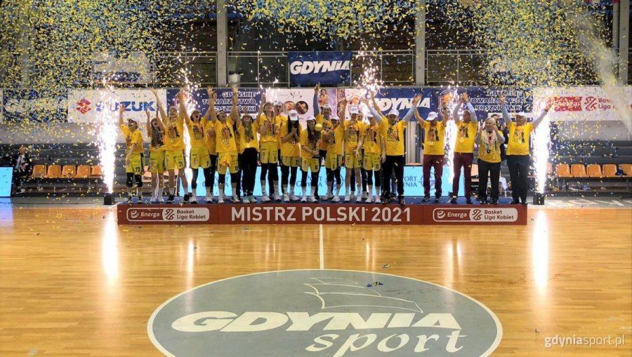 Koszykarki VBW Arki Gdynia podczas fety mistrzowskiej w 2021 roku. Cały zespół ustawiony jest w klubowych, żółtych strojach. Zawodniczki i sztab stoją w szeregu na podeście z podpisami "Mistrz Polski 2021" i Energa Basket Liga Kobiet. 