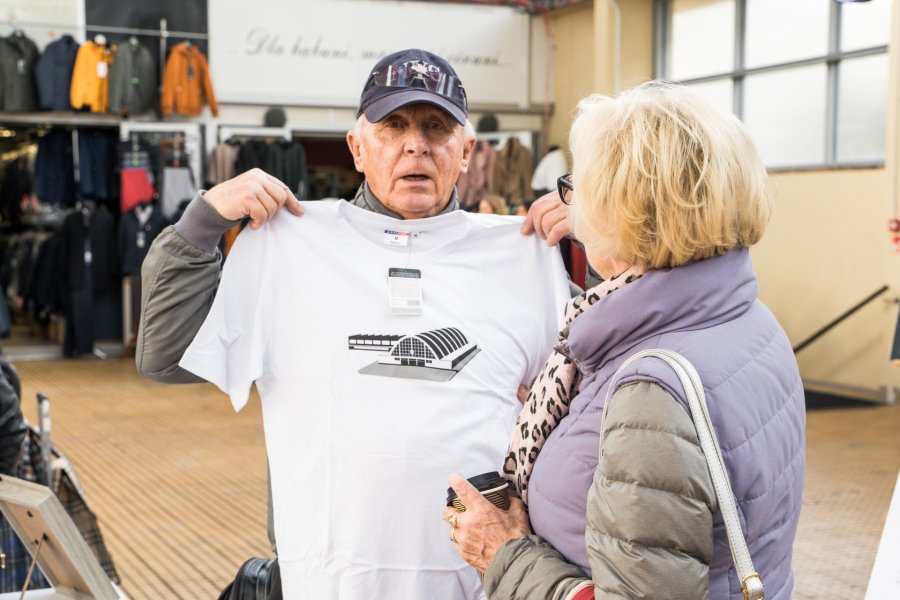 Starszy mężczyzna pozuje do zdjęcia z koszulką Hal targowych. Obok stoi tyłem do fotografa starsza kobieta.