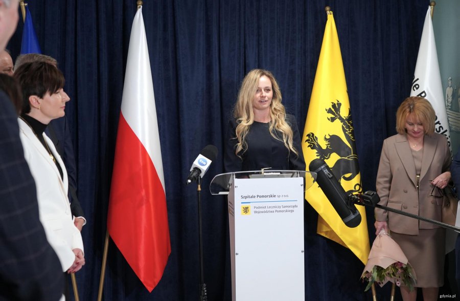 Kobieta z długmi blond włosami na m&oacute;winicy, obok flagi