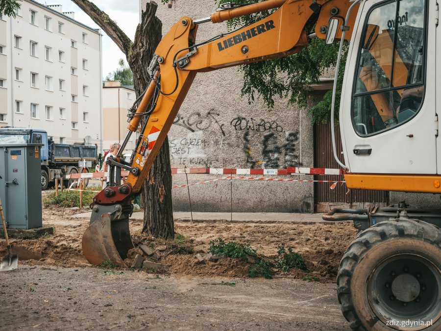 na pierwszym planie koparka wykonuje roboty drogowe, w tle prace drogowe