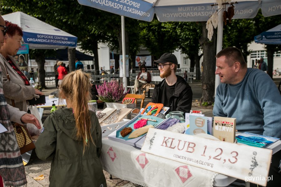 Stoisko gdyńskiego Klubu 1, 2, 3 na Festynie Tolerancji. Na pierwszym planie tabliczka z nazwą klubu. Za nią stolik wyłozony książkami i rękodziełem. Za stolikiem siedzi dwóch mężczyzn. Obok stoją przechodnie, którzy przyglądają się stoisku // fot. Karol Stańczak