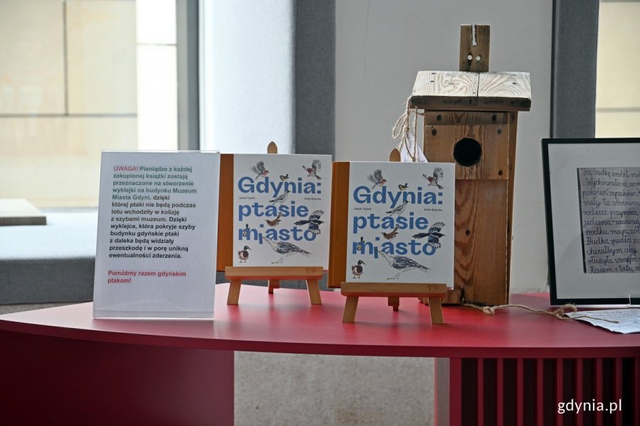 Dwa egzemplarze książki „Gdynia: ptasie miasto” oraz budka lęgowa na stoliku w holu Muzeum Miasta Gdyni. Po lewej - informacje, że dochód ze sprzedaży książki pokrył zakup i montaż wyklejki z markerami na szybach muzeum