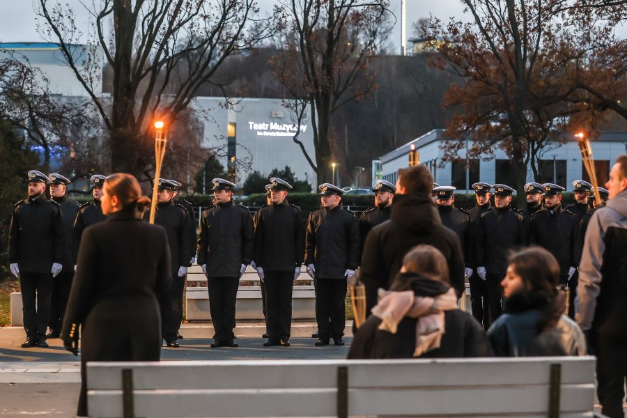 Na pierwszym planie: siedzące na ławce dziewczyny. Przed nimi stoją w rzędzie studenci z pochodniami. Po drugiej stronie stoi Kompania Reprezentacyjna Uniwersytetu Morskiego // fot. UMG