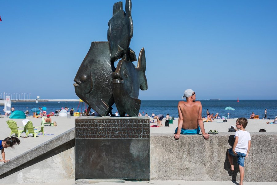 Pomnik - trzy ryby oraz tablica z napisem. W tle morze i plaża. 