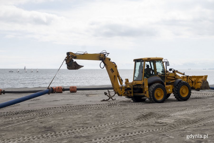 Żółta koparka ciągnie przywiązaną na lince rurę refulacyjną po piasku na plaży Śródmieście. W tle morze i żaglówki // fot. Sławomir Okoń