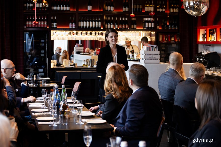 Kobieta przemawia stojąc w restauracji do ludzi siedzących przy stołach.