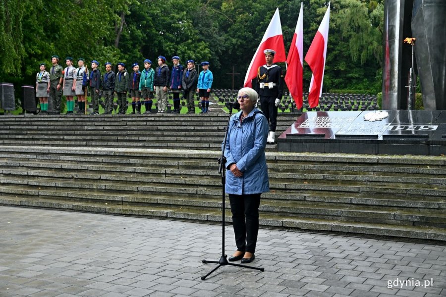 Joanna Zielińska - przewodnicząca Rady Miasta Gdyni przemawiająca podczas uroczystości z okazji 84. rocznicy agresji niemieckiej na Polskę, które odbyły się na Cmentarzu Wojennym w Redłowie. W tle posterunek honorowy przy pomniku Obrońców Wybrzeża, biało-czerwone flagi i harcerze ustawieni w szeregu 