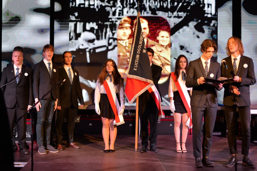 Na scenie z przodu deóch młodych mężczyzn, z tylu poczet sztandarowy, z lewej grupa uczniów, w tle na ekranie wyświetlone zdjęcie jednego z patronów