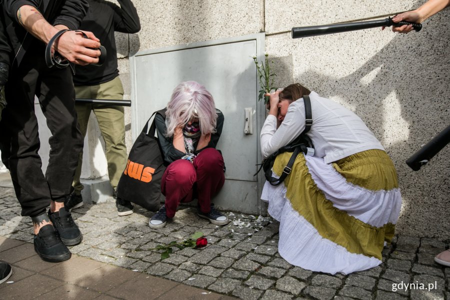 Fragment inscenizowanej akcji performatywnej zorganizowanej w ramach protestu "Stop przeciwko przemocy wobec narodu Białorusi". Dwie kobiety kulą się pod ścianą budynku. Nad nimi stoją funkcjonariusze z pałkami w rękach // fot. Karol Stańczak