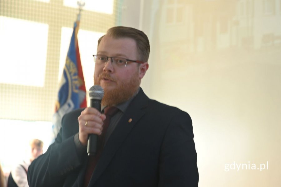 Stojący mężczyzna mówiący do mikrofonu w szkolnej hali sportowej