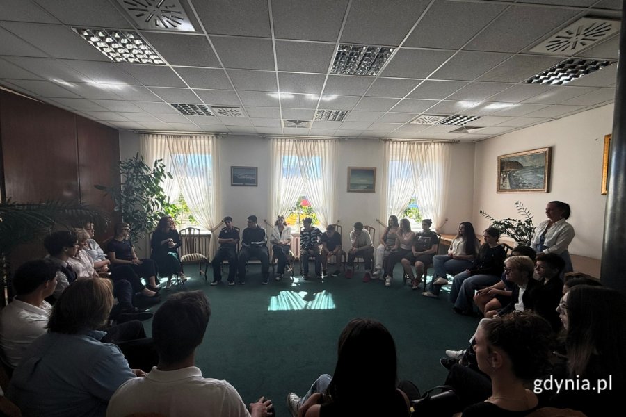 Szeroki kadr, sala wizytowa, a na niej przedstawiciele Młodzieżowej Rady Miasta Gdyni oraz młodzieży z Gruzji. Siedzą w okręgu na fotelach i rozmawiają.