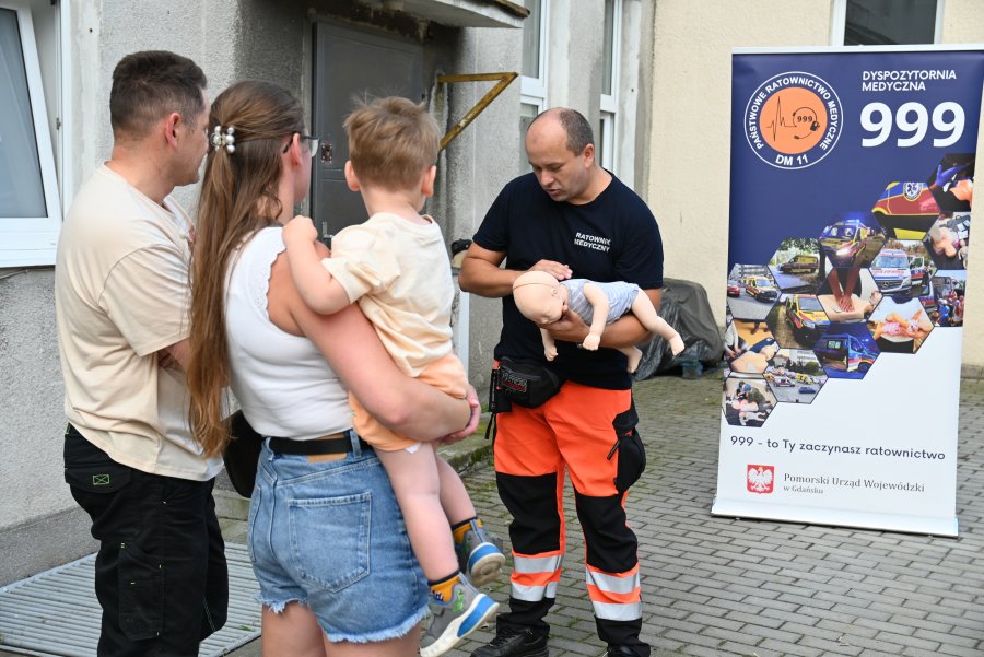 Para z małym dzieckiem przygląda się jak ratownik demonstruje pierwszą pomoc na manekinie wiekości dziecka
