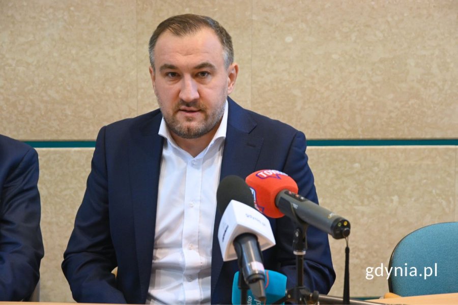 Mężczyzna w marynarce siedzący i mówiący do mikrofonu na tle beżowej ściany