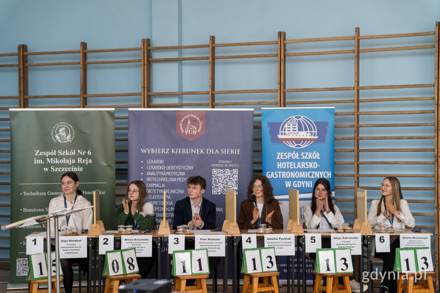 sześcioro uczestników konkursu, siedzą przy biurkach, w tle banery promocyjne i sala gimnastyczna 