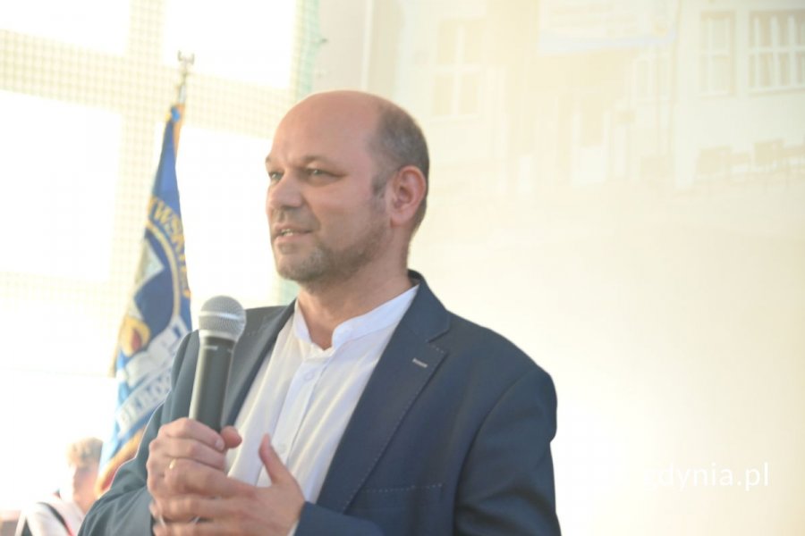 Stojący mężczyzna mówiący do mikrofonu w szkolnej hali sportowej