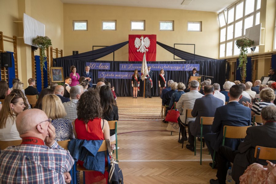 Sala, publiczność siedzi tyłem, z przodu ściana udekorowana udrapowanym granatowym materiałem, gosło Polski pośrodku, poniżej okolicznościowy napis