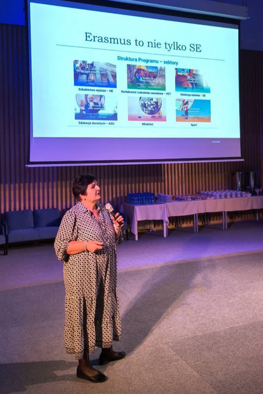 Kobieta przemawiajacy przed widownią z mikrofonem w ręku, wdalszym planie prezentacja