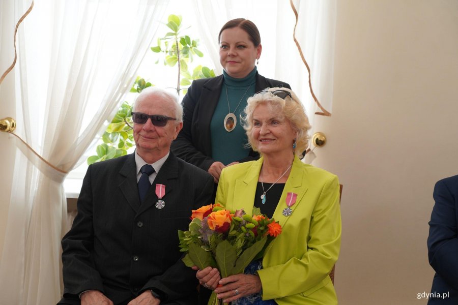 Para siedzi, kobieta trzyma pomarańczowe kwiaty, z tyłu stoi inna kobieta